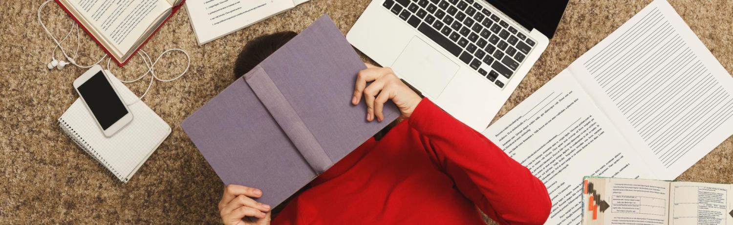 Student lying on the floor with a book over their face surrounded by papers, a laptop, phone and textbooks.