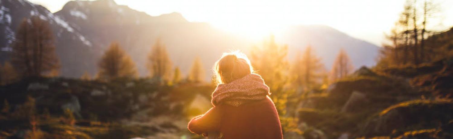 Girl sitting with her legs tucked in on a rock overlooking a mountain and sunset.