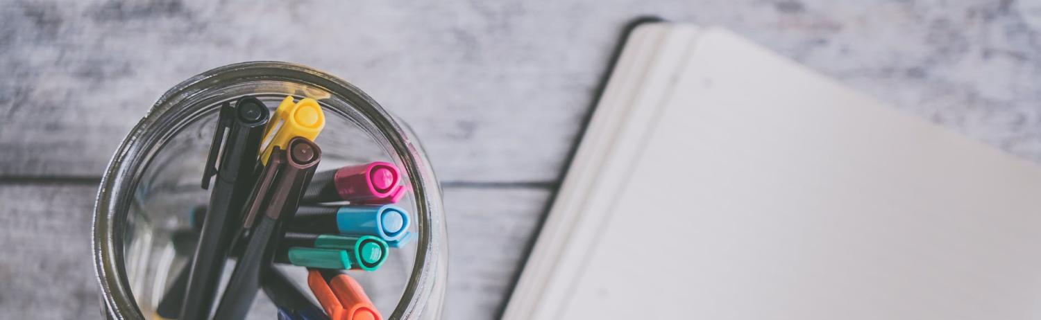 A mason jar of colorful pens sits next to a blank page of an open notebook.
