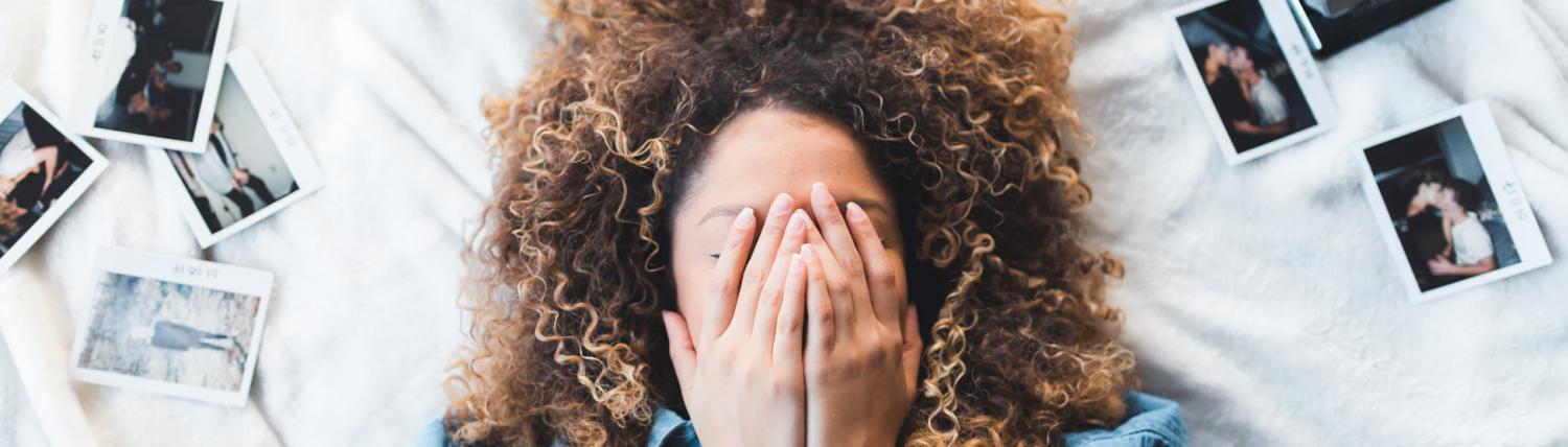 Girl with curly hair laying on her bed with her hands over her face surrounded by Polaroids.