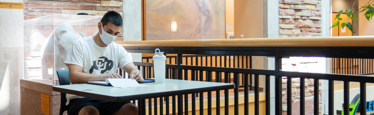 Student sitting at a desk in the UMC wearing a mask and CU Buff t-shirt as they flip through a notebook.