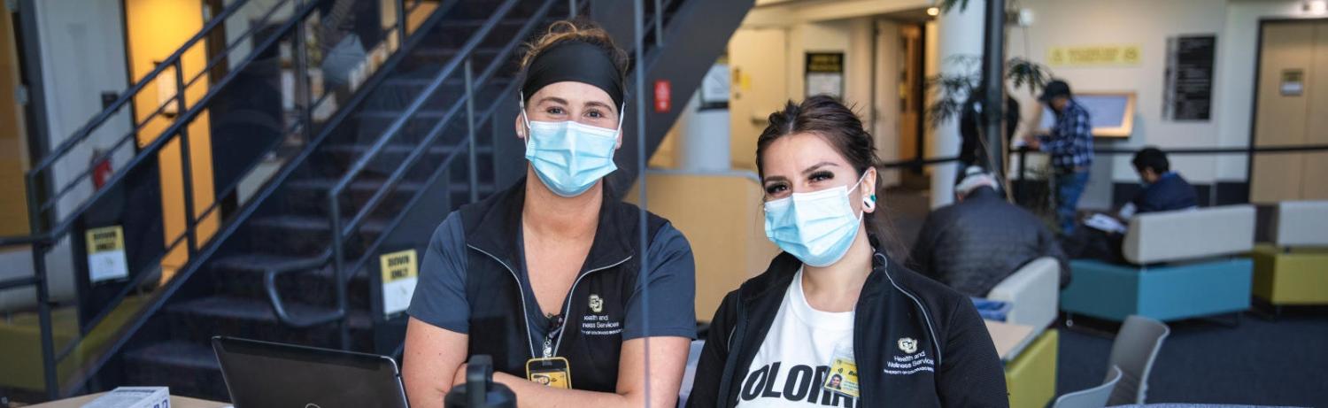 Two health care providers wearing masks sit at the check-in desk of the CU Boulder vaccination clinic site.