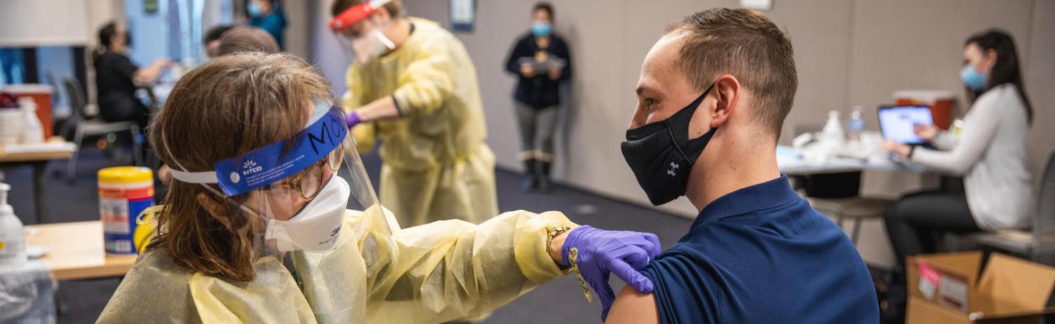 A nurse rolls up the sleeve of a masked man with short hair, preparing to give him a covid vaccine.