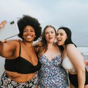 Photo of a group of friends with different body sizes taking a selfie on the beach in their bathing suits.