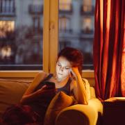 Girl sitting on her couch in the dark next to a lamp staring at her phone screen with a sad look on her face.