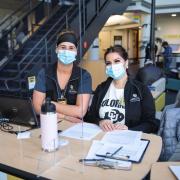 Two health care providers wearing masks sit at the check-in desk of the CU Boulder vaccination clinic site. 