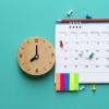 Wooden clock sitting next to a spiral calendar with colorful tacs.