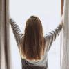 Girl standing in front of her window as she pulls back the blinds and lets the sunlight in. 