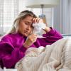 An ill looking girl holding a fistful of tissues to her forehead as she peers down at a thermometer.