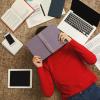 Student lying on the floor with a book over their face surrounded by papers, a laptop, phone and textbooks.