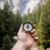 Hand holding a compass pointing north in front of a wooded background of pine trees.