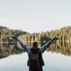 Girl wearing a beanie holding her arms up with joy in front of a forested lake.