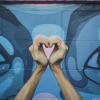 Person holding up their hands in a shape of a heart in front of a colorful outdoor mural.