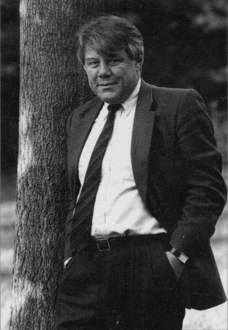 Thanasi Moulakis leans against a tree in black and white, wearing a suit and a striped tie, staring at the viewer, a half-smile on his lips