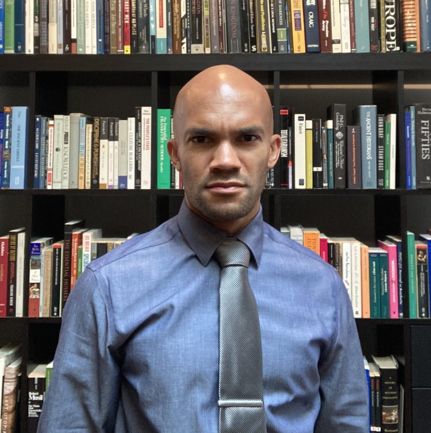 Photo of Michael Williams, an African American man wearing a blue dress shirt and gray tie standing in from of a bookcase