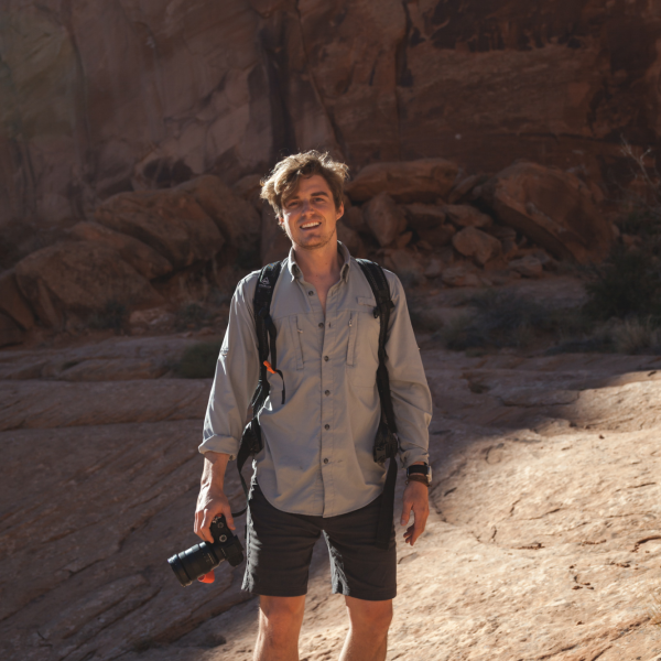 Thomas Balcom stands on a large boulder with a camera in his right hand.
