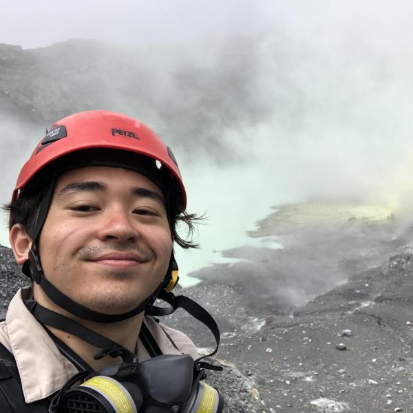 Justin Wang wears a hard hat and smiles from the edge of a volcano