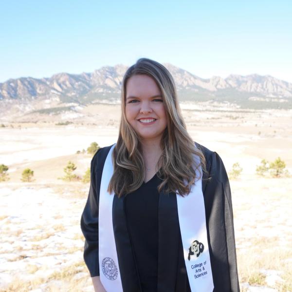 Kylie Conway wears her graduation gown and stands in a field of dry grass and snow with mountains in the background