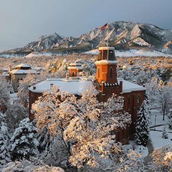 The rooftop of the Old Main building flies the American flag with snowy mountains in the background