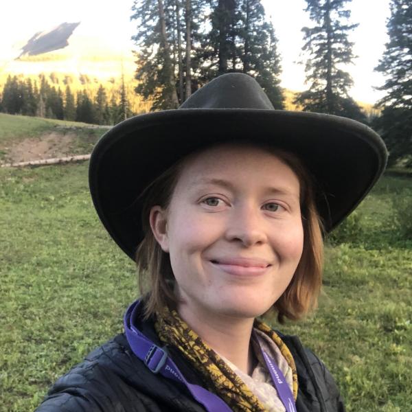 Shannon Ross wears a cowboy hat and stands in a grassy field with pine trees behind her
