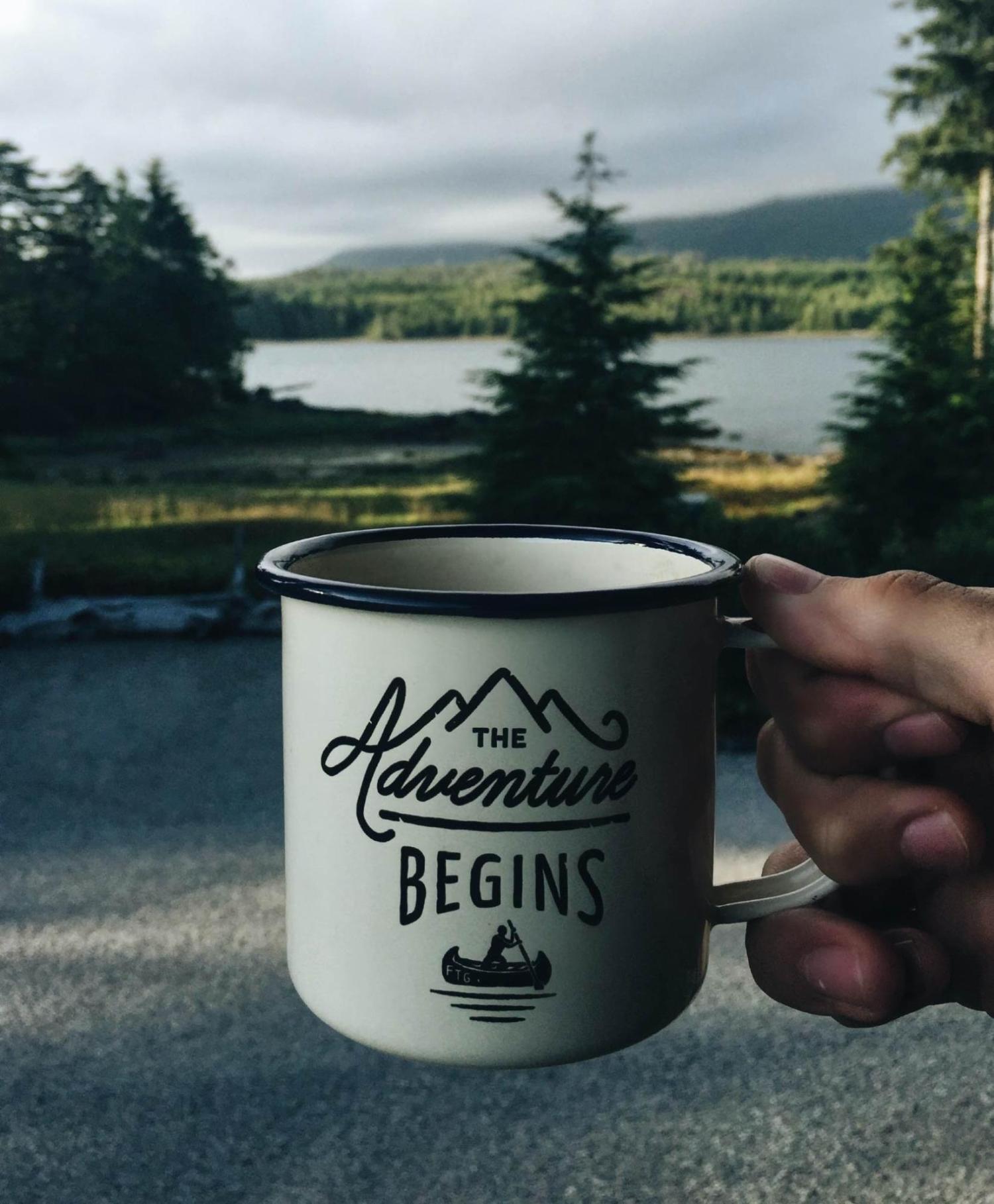 Hand holding coffee mug with "Adventure Begins" on it, with trees in the background