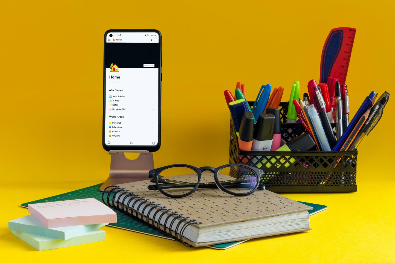 view of someones desk with a notebook, office supplies, glasses, and a smartphone, with a yellow background