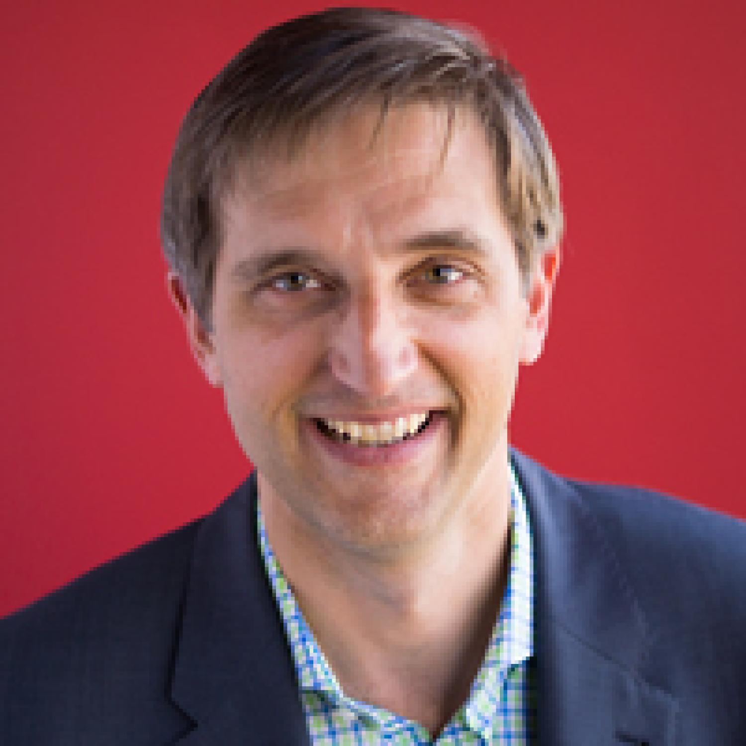 headshot of smilin man in a suit with a red backdrop