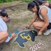 Three people crouch on a concrete surface, painting a black buffalo with 'CU' in gold. Colorful handprints and '2024' in pink chalk surround the design. They are outdoors, dressed in casual athletic clothing, and one person is smiling while painting.