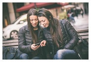 Adult female twins sitting on a bench looking at a cell phone. 