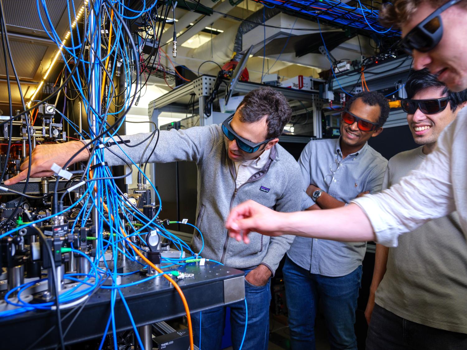 Researchers wearing safety glasses work with a complex setup of wires and equipment in a high-tech lab, examining and discussing the system.