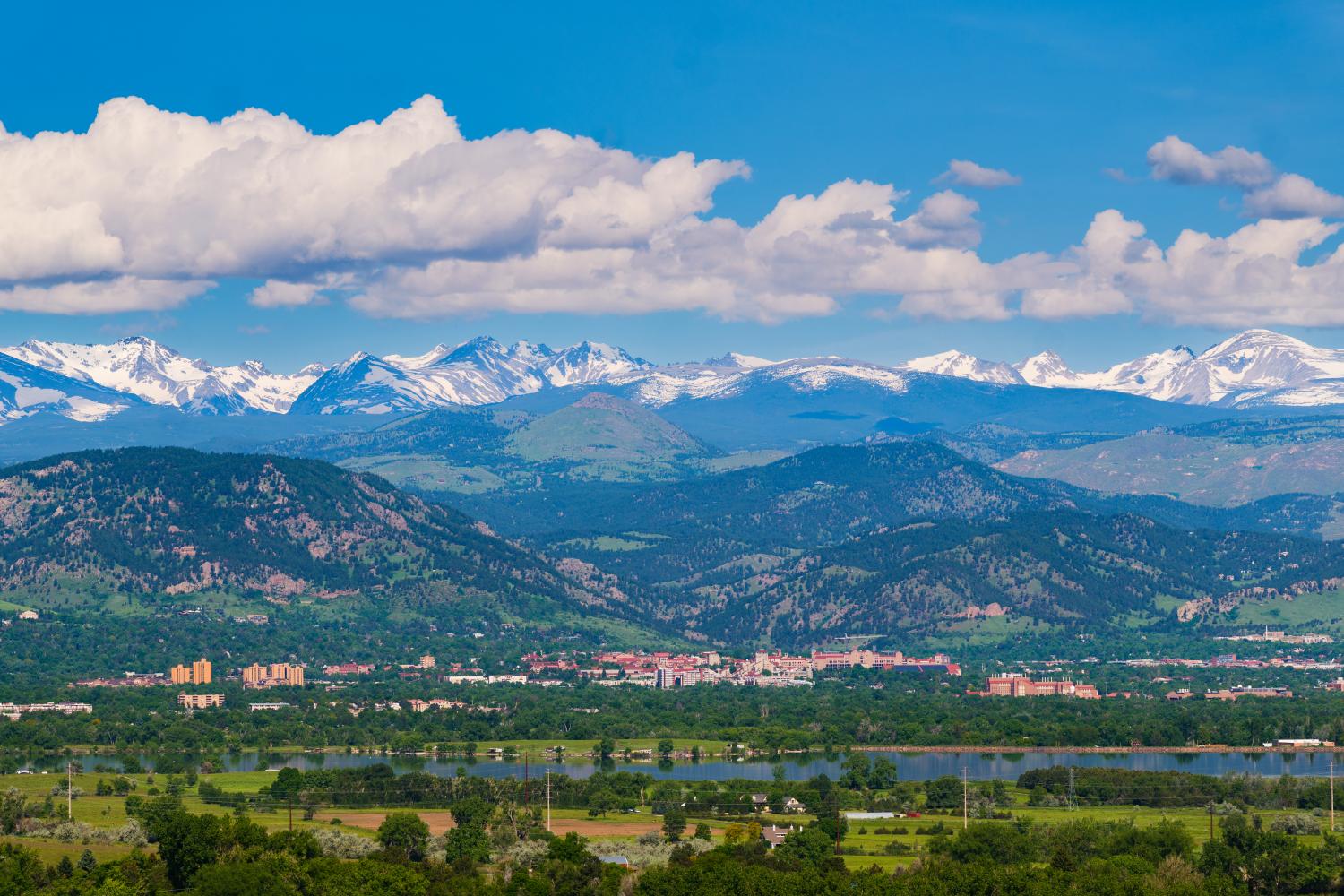 A full landscape of the city of Boulder that includes CU Boulder campus buildings, many layers of mountains, cloudy blue skies, and lush greenery.