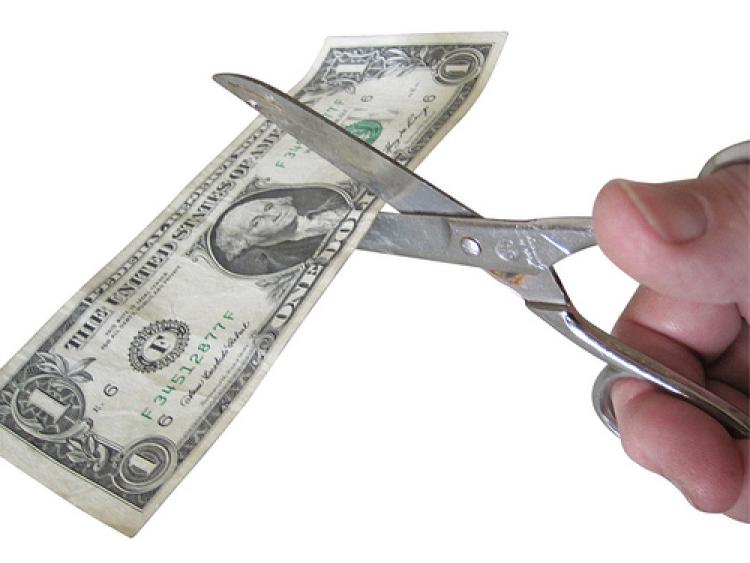 A hand holding scissors cutting a one dollar bill.(© by Images of Money/Flickr Creative Commons)