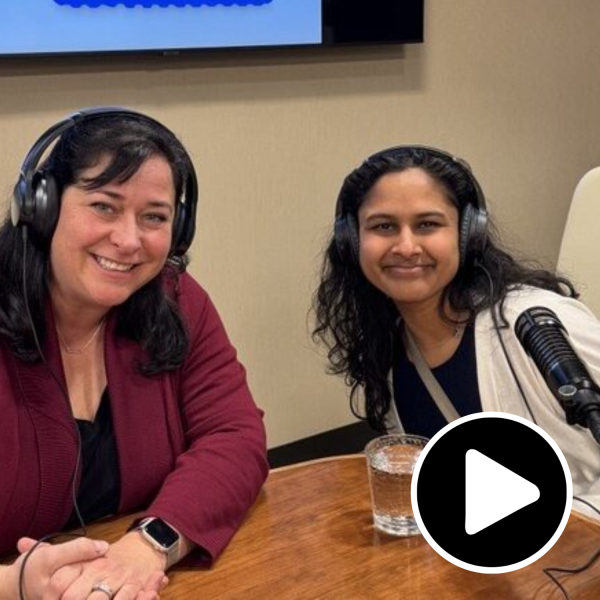 A white woman and a woman of color sit at a desk with headphones and microphones for reporting a podcast and smile at the camera