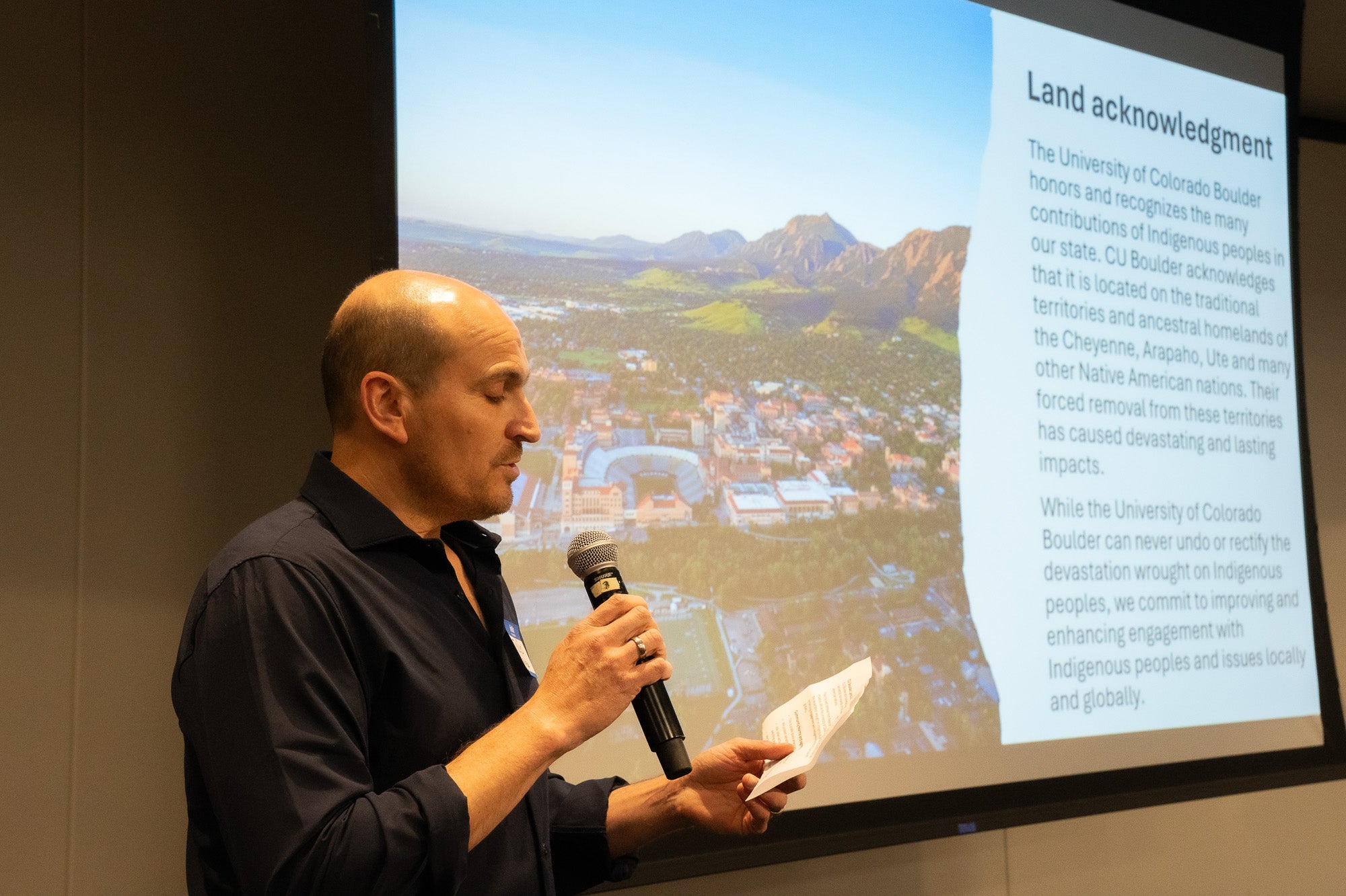 A man reads from a paper into a microphone in front of a projector screen
