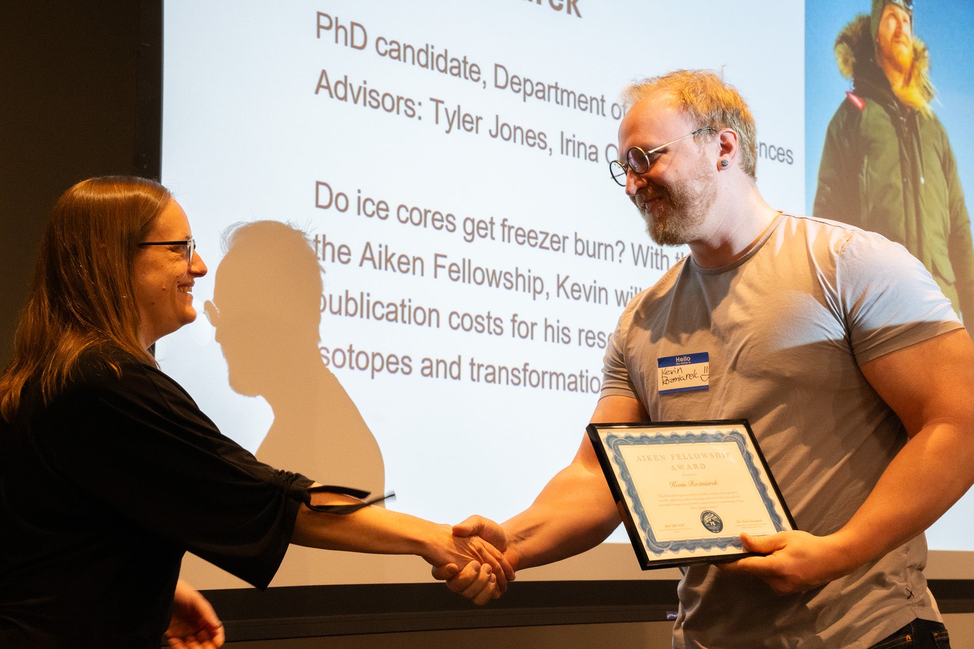 A man in wire-framed glasses and a t-shirt holds a plaque with his left hand while he shakes the hand of a woman in black with glasses in front of a projector screen
