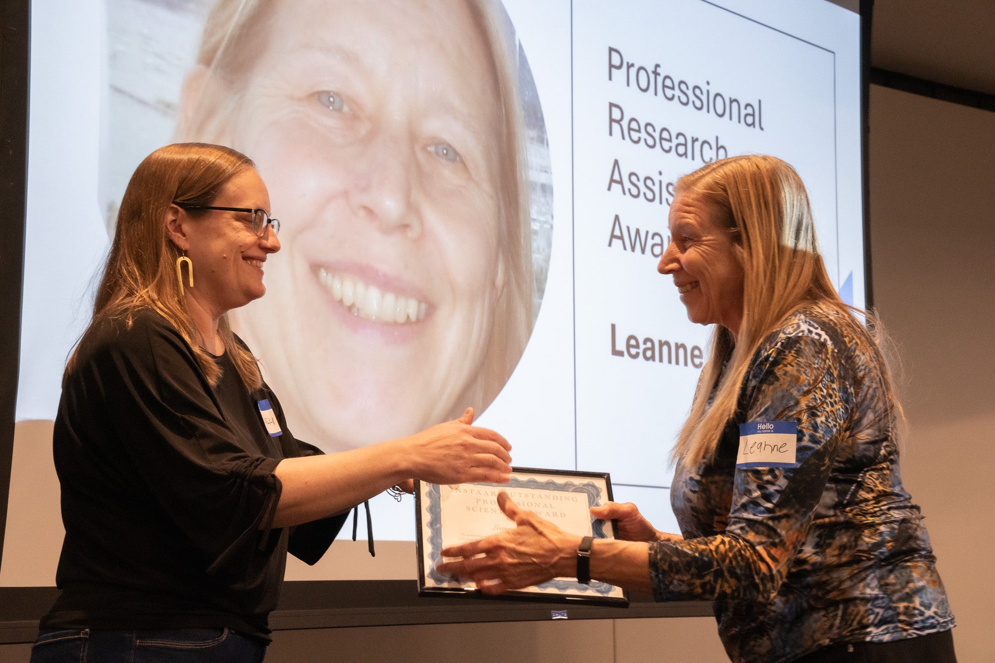 Two women shake hands and hand off a plaque in front of a projector screen