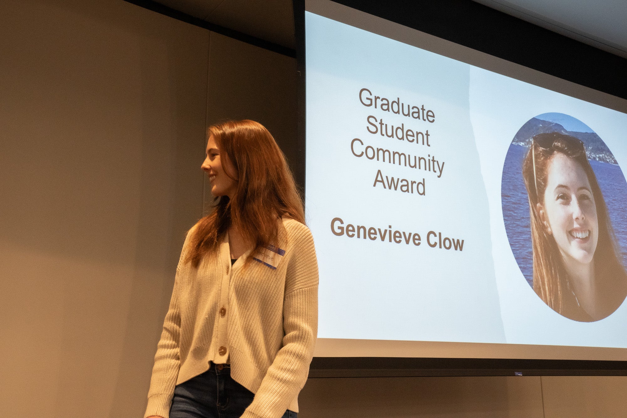 A young woman looks out of frame in front of a projector screen with a photo of her face that reads "Graduate Student Community Award Genevieve Clow"