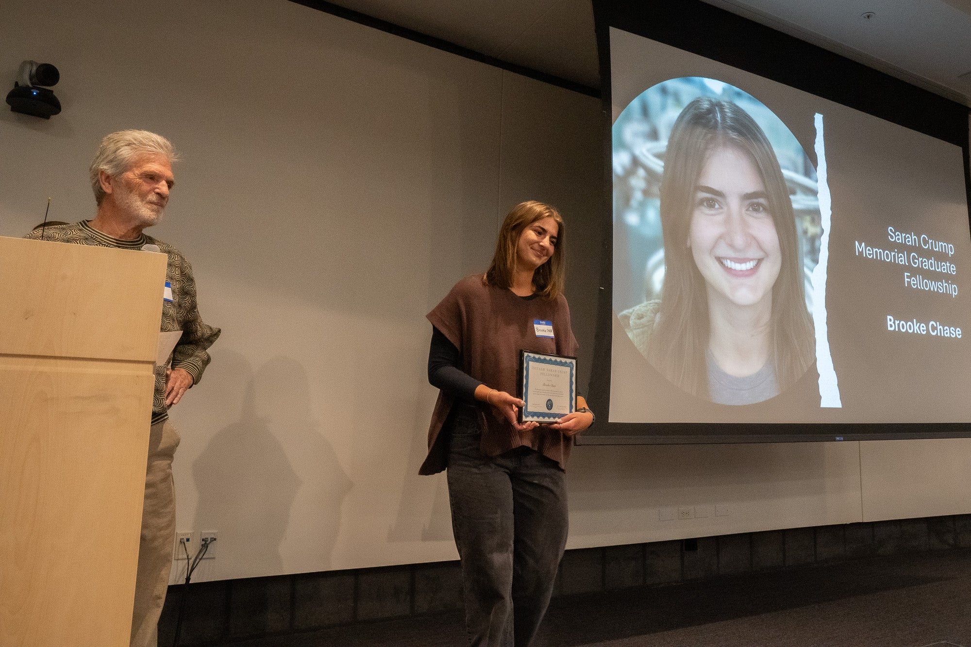A young woman stands holding a plaque center stage. To the right is a projector screen that reads "Sarah Crump Memorial Graduate Fellowship: Brooke Chase," to the left an older man stands behind a podium