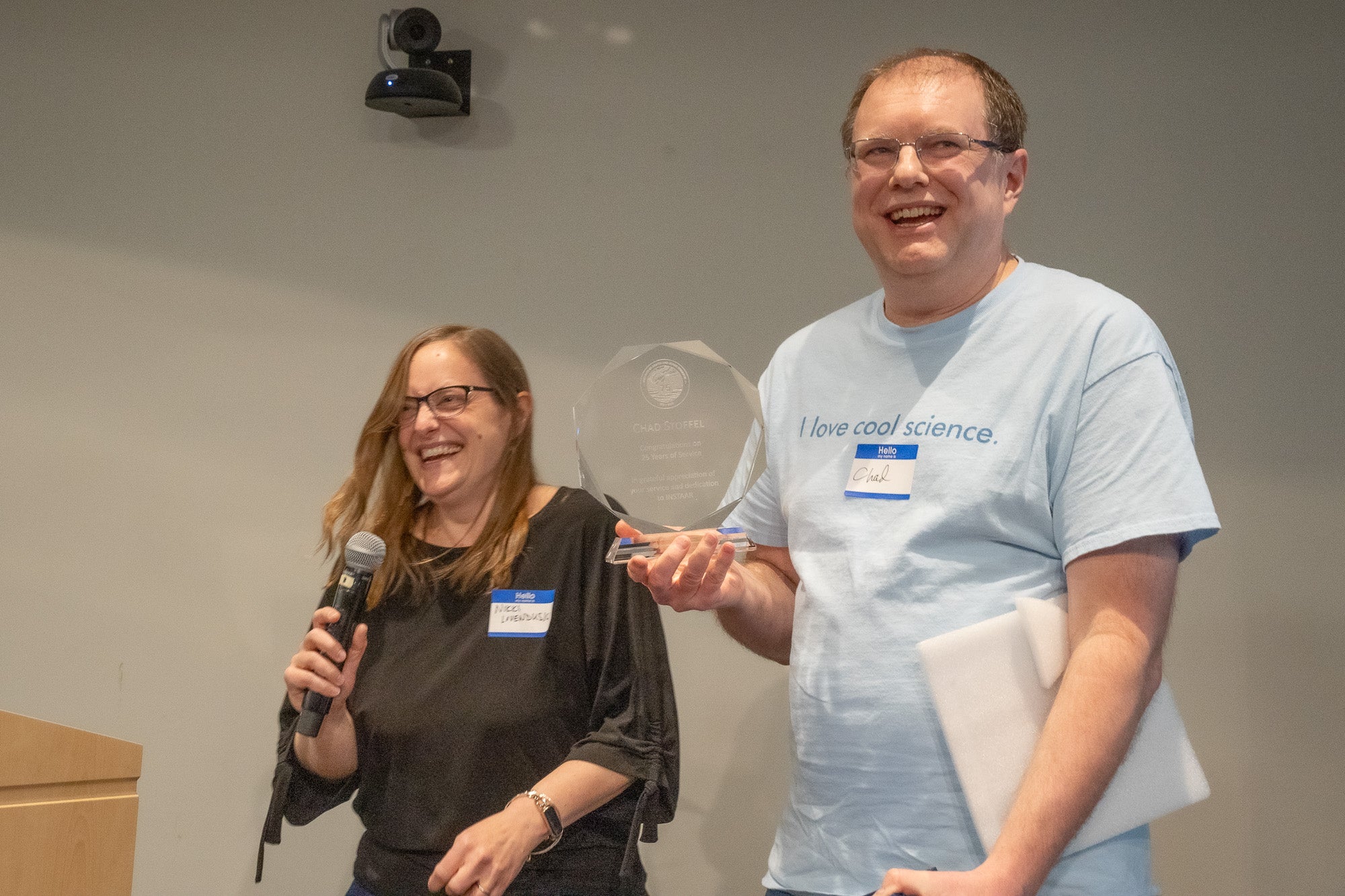 A man in a blue t shirt holds up a glass trophy next to a woman in black with a microphone. Both are smiling and laughing.