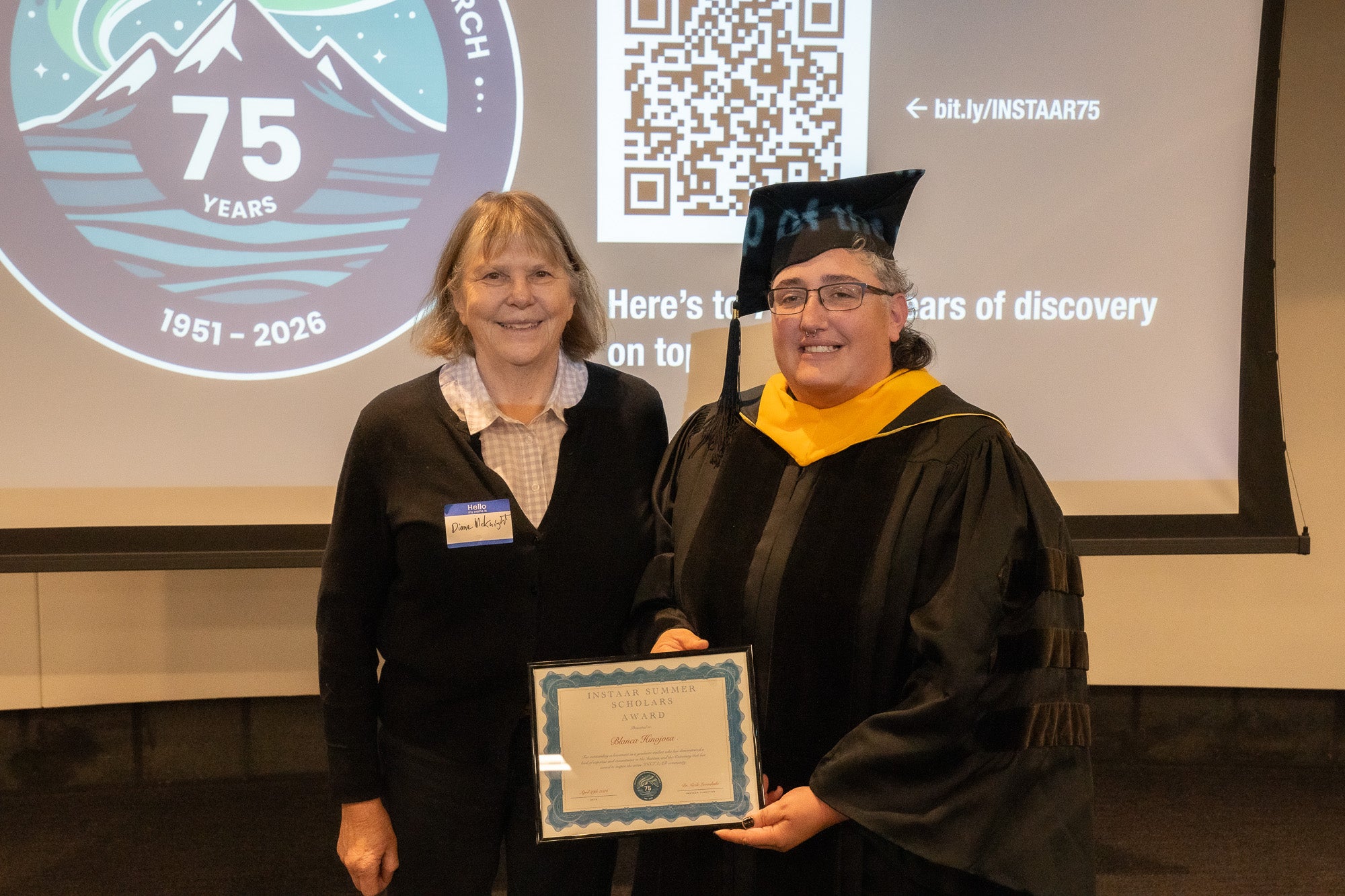 A woman in a button down and cardigan poses with a woman wearing a tassel, robe and cap and holding a plaque in front of a projector screen