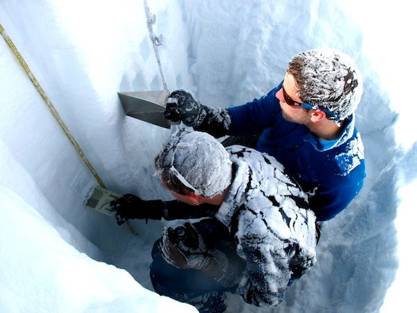 Two hydrology researchers, with snow-covered hair and backs, take snow samples from the sidewall of a snow pit