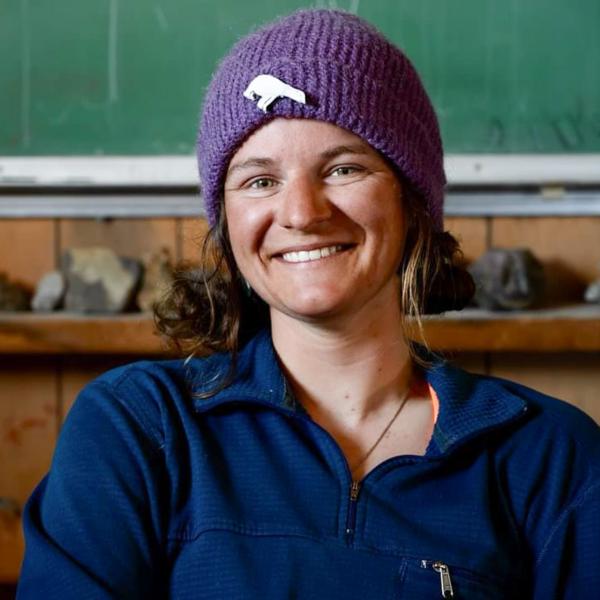 A young brunette woman in athletic wear smiles for the camera while seated in front of a rock collection and a green chalkboard