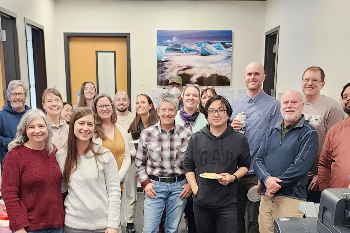 A group of about 20 INSTAAR folks, including front office staff, pose for a photo in the INSTAAR front office. A copier machine partially obstructs the photo.