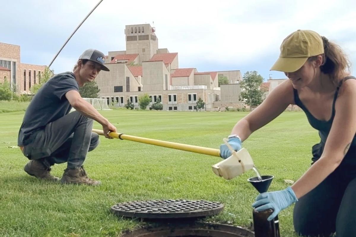 Squatting next to an opened manhole cover on a green campus lawn, a research partner holds a long pole with an attached water sample cup and tips it into a funnelled container held by a rubber-glove wearing Alli Cook. This is one of many water samples that Alli gathered from CU Boulder storm sewers.