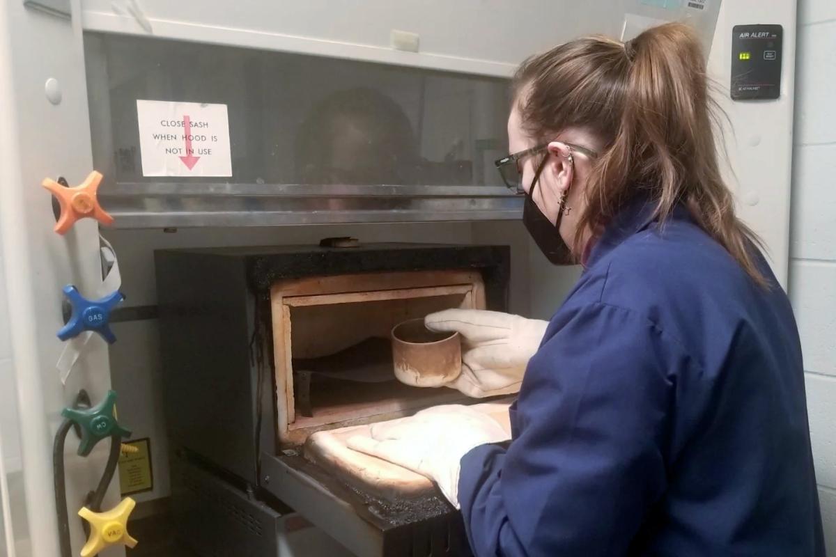 Grad student Mackenzie Bowden, wearing thick mitts, places a sample of an urban material, like PVC pipes, into a lab oven where it will be combusted under controlled conditions and its ash leached into water and measurements made