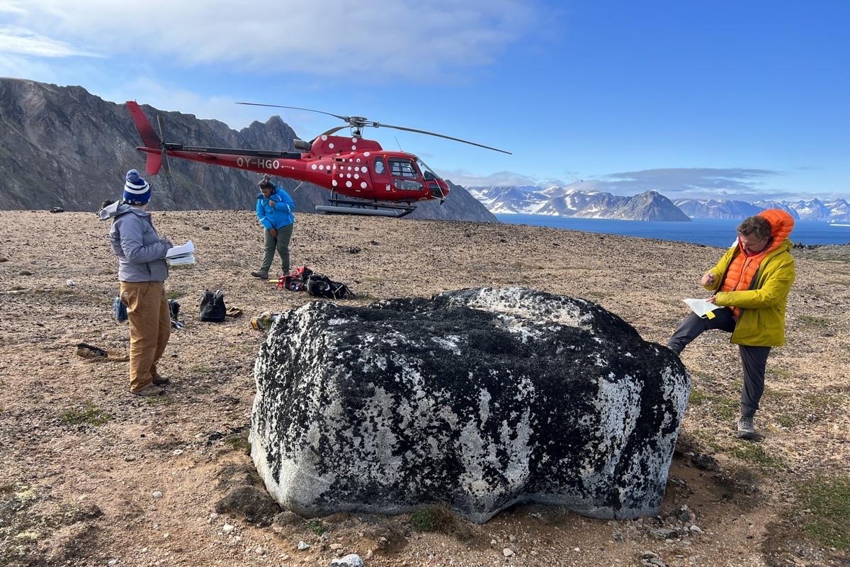 Three researchers gather around a very large boulder on a flat surface high above the ocean. A red helicopter is behind them. In the distance are rugged mountains and fjords.
