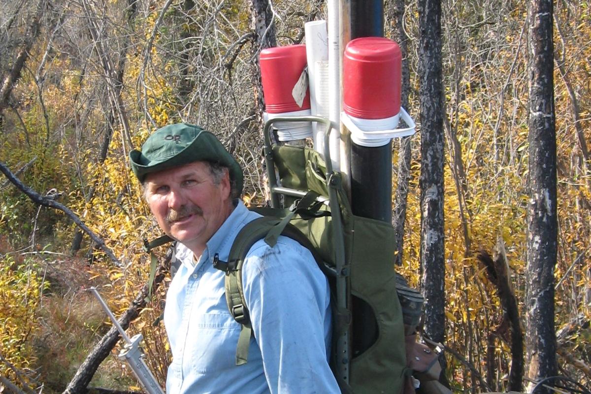 George Aiken gears up for fieldwork, wearing a backpack with long tubes and instruments, and surrounded by other equipment