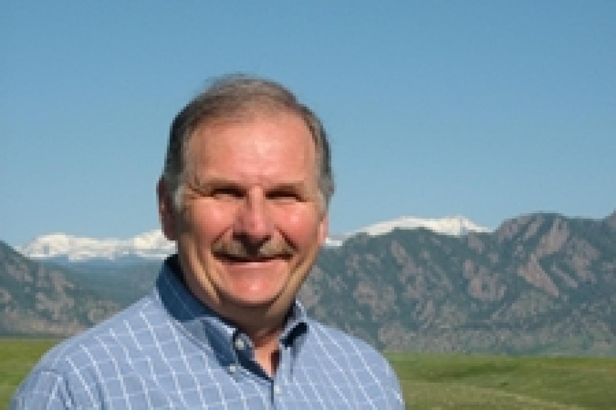 George Aiken, in blue button shirt, stands in sunshine and a grassy field, with the Colorado Front Range in background.