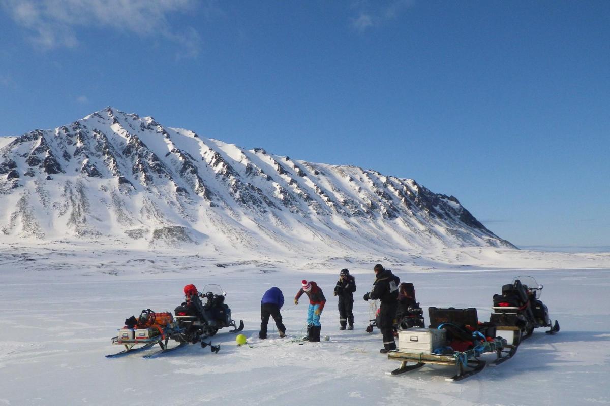Four bundled-up researchers park their snowmobiles on a frozen lake with a jagged mountain in the background