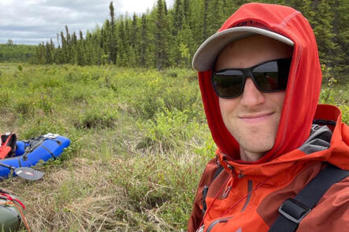 Dylan Blaskey, in red hooded jacket and gray waders, works near an arctic river, with grasses and conifers in the background